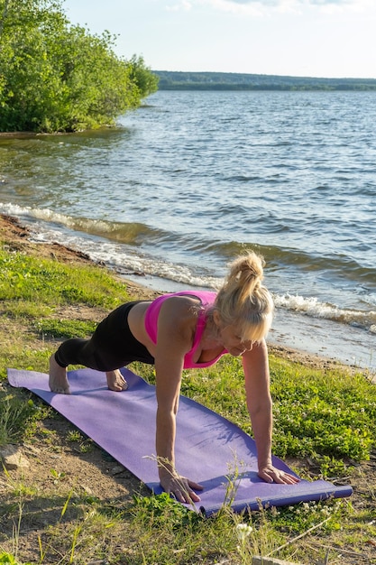 Una donna anziana in abbigliamento sportivo in piedi su una tavola che pratica yoga sulla riva del lago Yoga nel parco