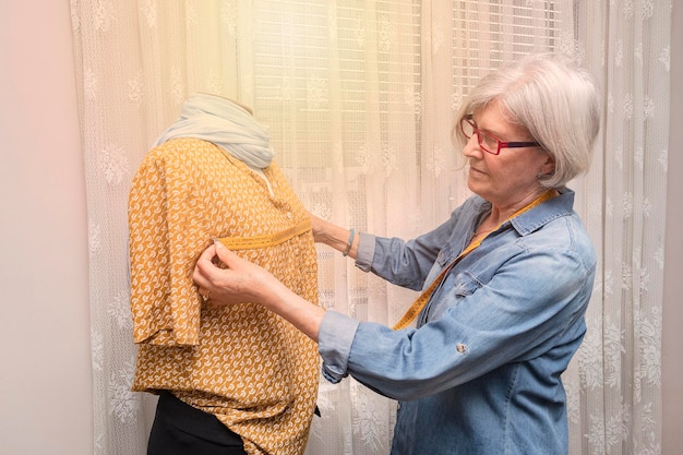 Una donna anziana misura con un metro a nastro il busto di un manichino vestito con una blusa arancione