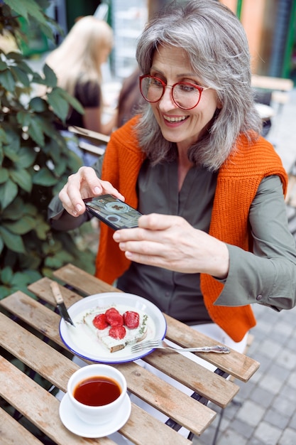 Una donna anziana positiva scatta una foto di un delizioso dessert alla fragola sulla terrazza del caffè all'aperto