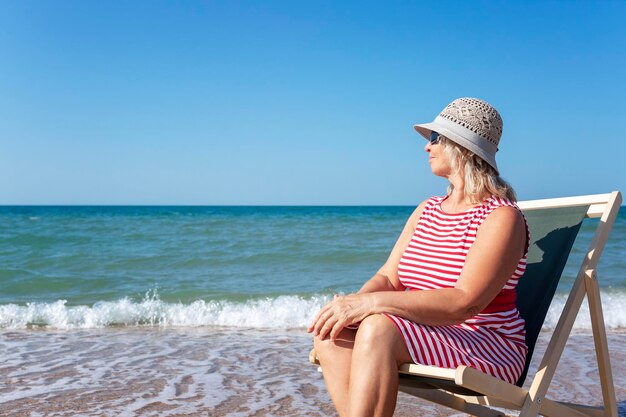 Una donna anziana sorridente con un vestito a righe si siede su un lettino sulla riva del mare in una giornata di sole. Rilassamento e tranquillità. Stile di vita attivo, energia e salute.