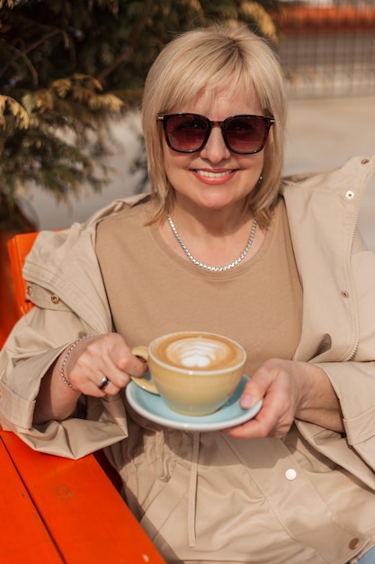 Una donna anziana sorridente felice con occhiali da sole alla moda in elegante abito beige si siede in una soleggiata giornata primaverile all'aperto e beve caffè