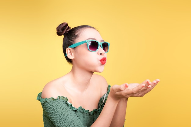 Una donna asiatica alla moda con occhiali da sole alla moda manda un bacio su uno sfondo giallo brillante