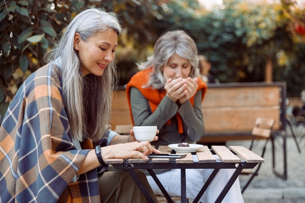 Una donna asiatica anziana usa il cellulare mentre un amico canuto beve il tè in un caffè di strada