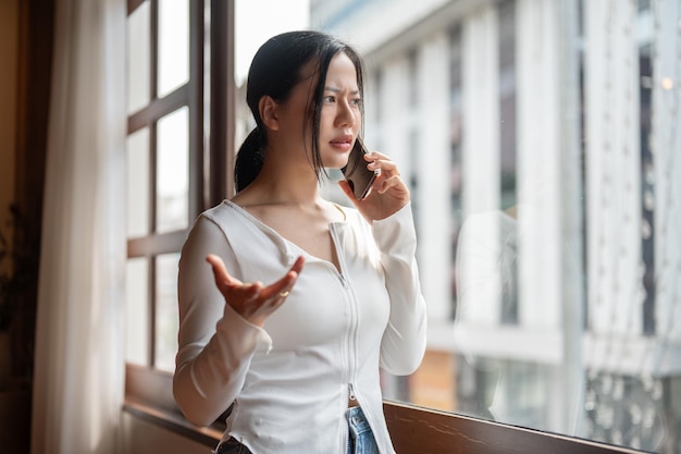 Una donna asiatica arrabbiata e insoddisfatta sta parlando al telefono stando in piedi vicino alla finestra