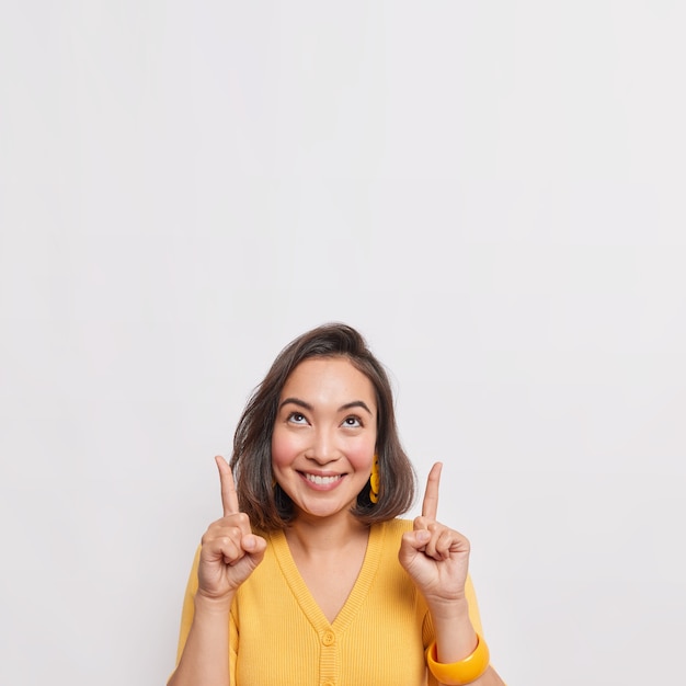 Una donna asiatica bruna positiva indica sopra e sorride piacevolmente indossa orecchini e bracciale gialli a ponticello isolati su un muro bianco