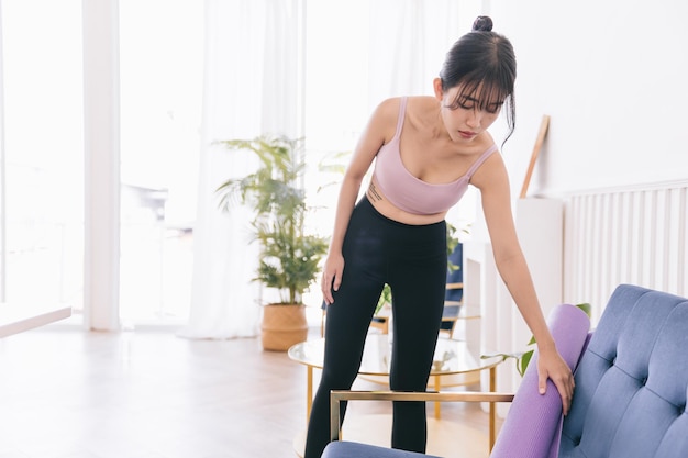 Una donna asiatica che indossa abbigliamento sportivo e pantaloni da yoga prende un tappetino da yoga arrotolato dalla sedia Allenarsi a casa Abitudini sane mantenersi in forma concetti di perdita di peso