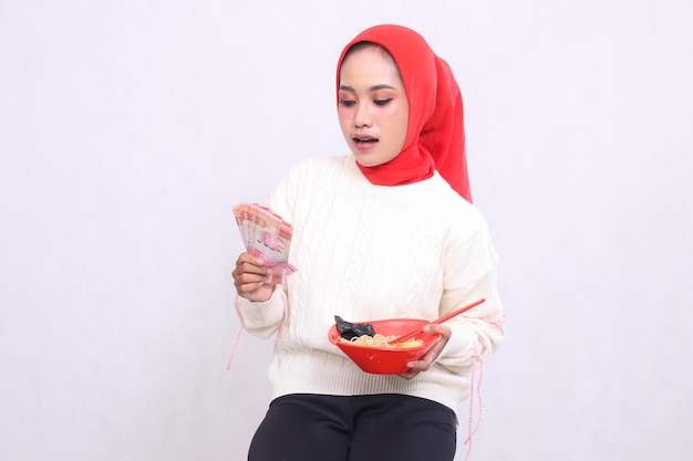 Una donna asiatica che indossa un hijab è scioccata nel vedere banconote di rupiah e una ciotola contenente ramen di cibo cinese