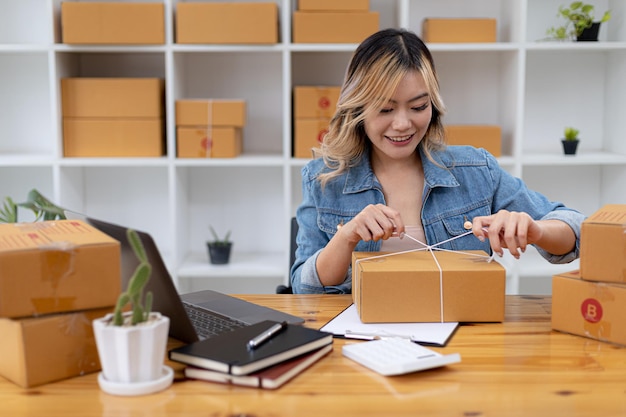 Una donna asiatica che lega un pacco alla scatola di un cliente, possiede un negozio online, fa le valigie e spedisce tramite una compagnia di trasporti privata. Concetti di vendita online e shopping online.