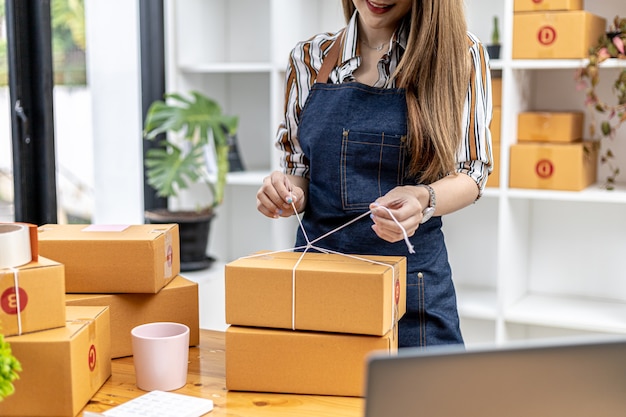 Una donna asiatica che lega un pacco alla scatola di un cliente, possiede un negozio online, imballa e spedisce tramite una compagnia di trasporti privata. Concetti di vendita online e shopping online.