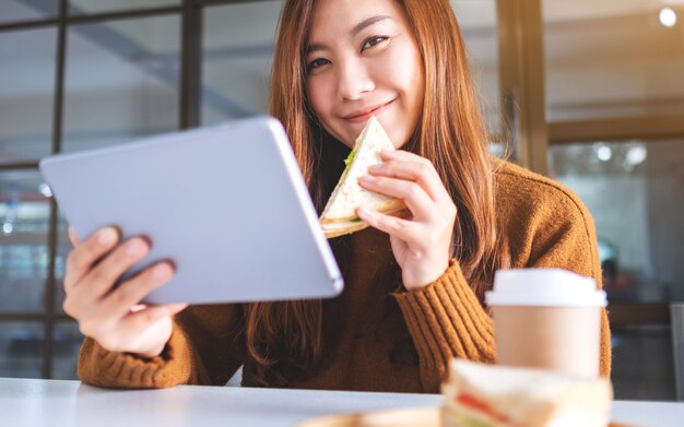 Una donna asiatica che tiene e mangia un panino integrale mentre usa un tablet pc