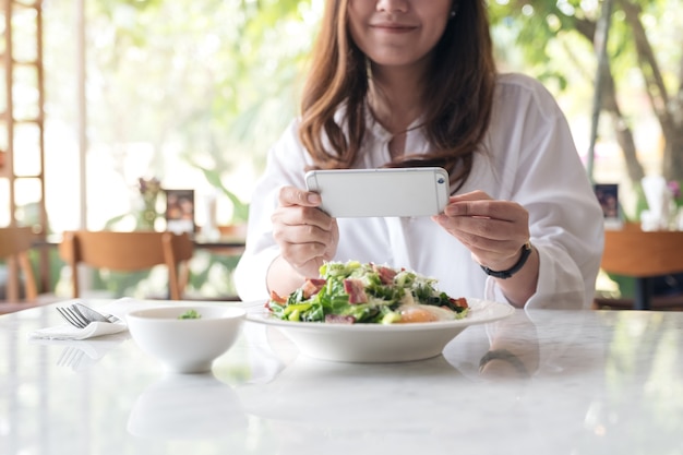 una donna asiatica che utilizza smartphone per scattare foto di insalata in un piatto bianco sul tavolo