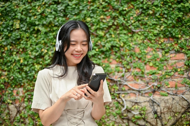 Una donna asiatica felice sta mandando messaggi con i suoi amici e ascolta musica mentre si rilassa su una panchina