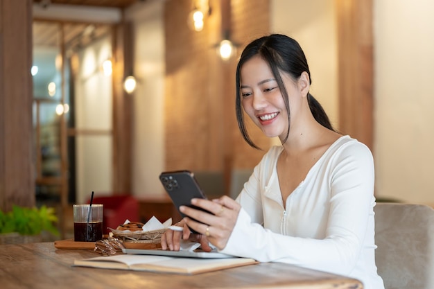 Una donna asiatica felice sta usando il suo smartphone per mandare messaggi ai suoi amici mentre è seduta in un bar