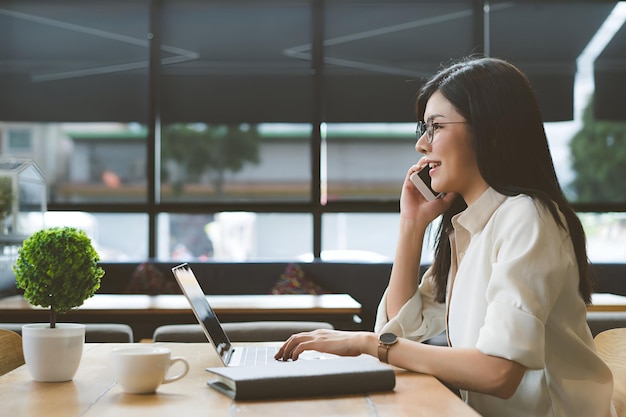 Una donna asiatica freelance che parla con lo smartphone del suo lavoro usando un computer portatile digitale che beve caffè a colazione sul tavolo del posto di lavoro in un caffè