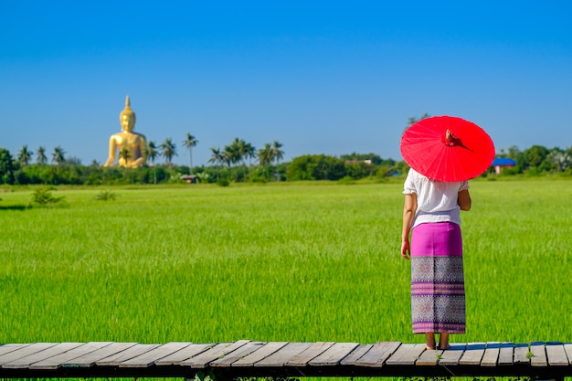 Una donna asiatica in possesso di un ombrello rosso che cammina su un ponte di legno nel campo di riso con una grande immagine dorata del Buddha.