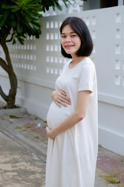 Una donna asiatica incinta che indossa abiti casual bianchi è in piedi sorridente di buon umore all'aperto.