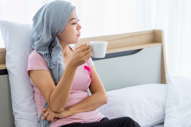 Una donna asiatica malata di cancro mammario che tiene una tazza di caffè con nastro rosa che indossa il velo Dopo il trattamento alla chemioterapia a letto Nella camera da letto a casa, assistenza sanitaria, medicina