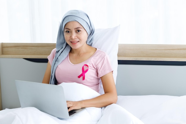 Una donna asiatica malata di cancro mammario con nastro rosa che indossa il velo Dopo il trattamento alla chemioterapia con attività lavorativa al computer portatile sul letto Nella camera da letto presso la medicina domestica