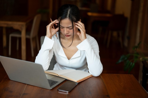 Una donna asiatica stressata che lavora da remoto in un bar e soffre di mal di testa mentre legge un libro
