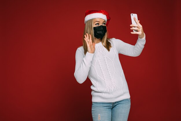 Una donna attenta con cappello da Babbo Natale e maschera protettiva chiama i suoi amici mentre trascorre il Natale in isolamento. Copia spazio