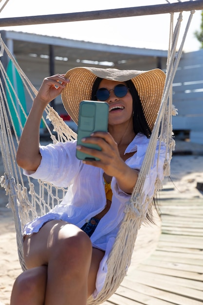 Una donna bi-razziale felice con gli occhiali da sole e il cappello da sole seduta in una amaca a fare selfie sulla spiaggia soleggiata.