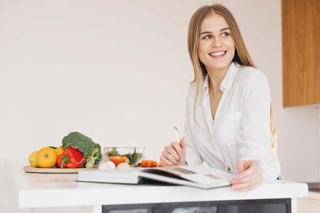 Una donna bionda carina e felice sta scrivendo qualcosa e sta guardando un libro di ricette in cucina