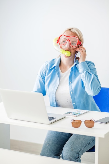 Una donna bionda con una maschera da sub si siede in ufficio e annuncia felicemente per telefono che sta andando in viaggio o sta comunicando con un tour operator Foto verticale