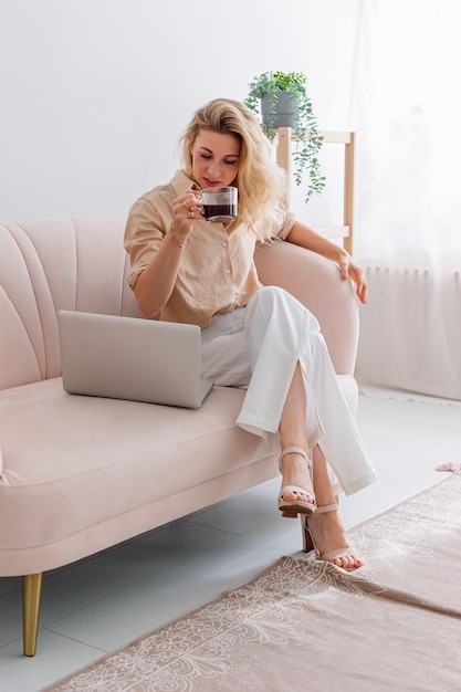 Una donna bionda d'affari che lavora al computer portatile si siede sul divano con una tazza di caffè