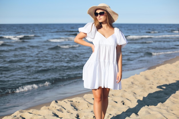 Una donna bionda felice e sorridente sta posando sulla spiaggia dell'oceano con occhiali da sole e un cappello