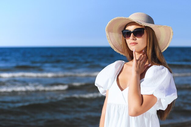 Una donna bionda felice sta posando sulla spiaggia dell'oceano con gli occhiali da sole e un cappello.
