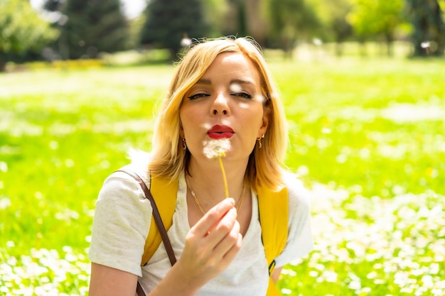 Una donna bionda sorridente che soffia sulla pianta del dente di leone che indossa un cappello e occhiali da sole seduta sull'erba in primavera accanto alle margherite in un parco della città