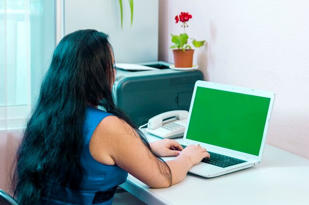 Una donna bruna in un ufficio a casa lavora con un laptop alla telecamera con schermo verde. Foto orizzontale