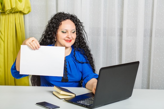 Una donna bruna riccia si siede a un tavolo in ufficio, tiene un foglio di carta e digita sul computer. Foto orizzontale