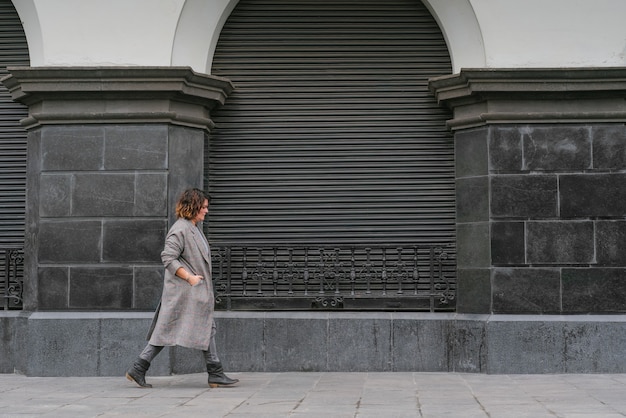 Una donna cammina per una vecchia strada guardando il pavimento e tiene le mani dentro il lungo cappotto, sullo sfondo di una vecchia architettura in scala di grigi. Copia spazio