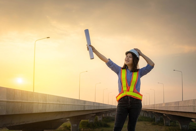 Una donna capo architetto o ingegnere sta alla costruzione del ponte indossando un elmo e lancia un progetto per esprimere la sua gioia quando il progetto è completo