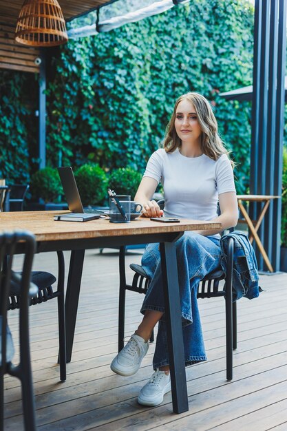 Una donna carina beve caffè sulla terrazza di un caffè estivo e lavora su un portatile a distanza mentre è in vacanza in un caffè con un portatile