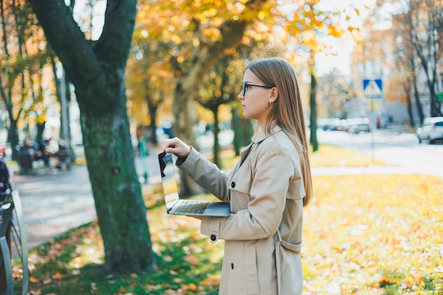 Una donna carina con gli occhiali e i capelli lunghi cammina in un parco autunnale con un laptop in mano Lavora online all'aria aperta
