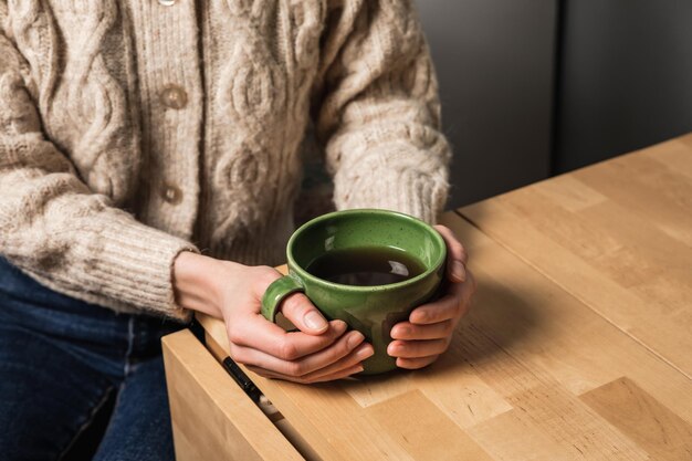 Una donna caucasica in un accogliente cardigan beige con in mano una tazza di tè verde seduta al tavolo