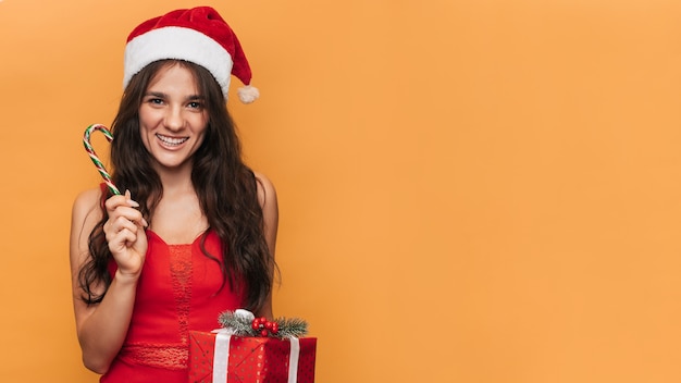 Una donna caucasica sorridente con un cappello da Babbo Natale rosso con una confezione regalo in mano su uno sfondo giallo isolato. Il concetto di Natale e Capodanno. Un posto per il tuo testo.