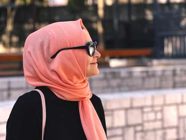 Una donna che indossa un hijab rosa e una camicia nera si affaccia su una strada cittadina.