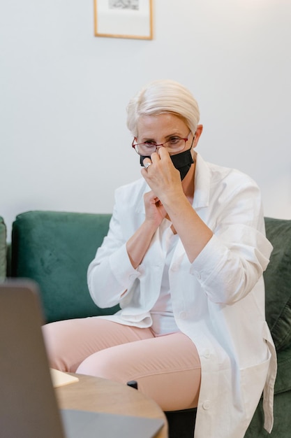 Una donna che indossa una maschera è seduta su un divano davanti a un laptop.