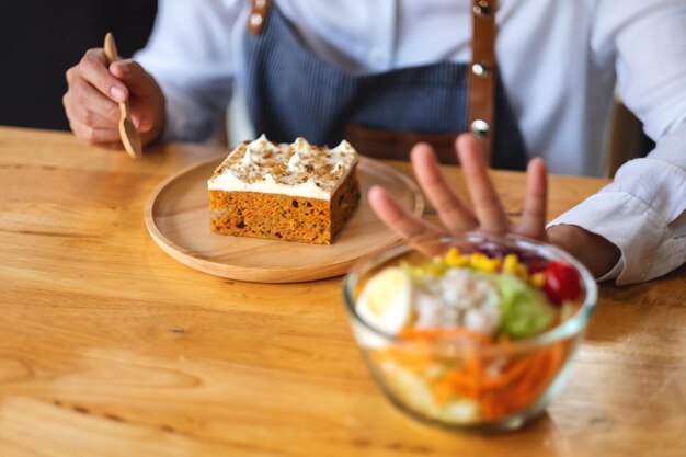 Una donna che sceglie di mangiare una torta e fa segno con la mano di rifiutare un'insalata di verdure sul tavolo