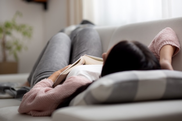 Una donna che si addormenta durante il giorno su un divano mentre cerca di leggere un libro.