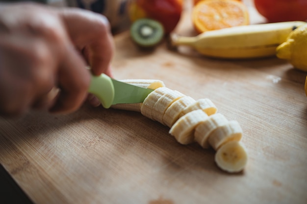 Una donna che taglia una banana con un coltello verde su un tavolo di legno circondato da altri frutti.