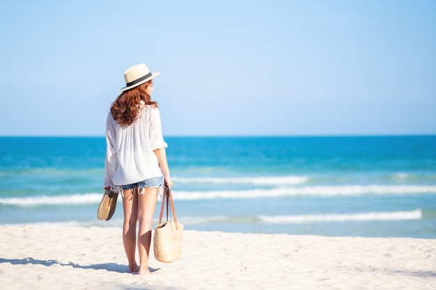 Una donna che tiene borsa e scarpe mentre passeggia sulla spiaggia con il mare e il cielo azzurro sullo sfondo