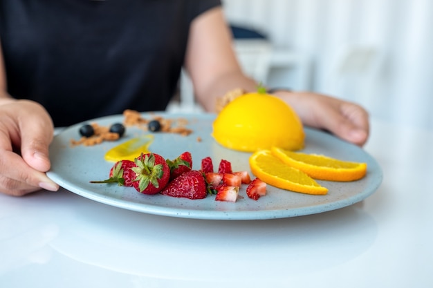 Una donna che tiene e mostra un piatto di torta all'arancia con frutta mista nella caffetteria