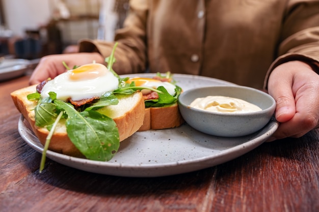 Una donna che tiene un piatto di panino per la colazione con uova, pancetta e panna acida sul tavolo di legno