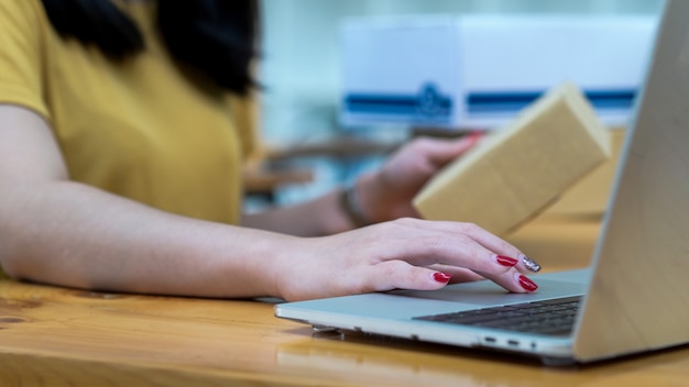 Una donna che vende online utilizza un laptop e tiene in mano una cassetta dei pacchi per controllare le informazioni sui clienti.