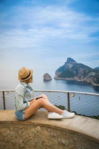 Una donna con cappello di paglia seduta sul bordo del Mirador de El Colomer godendo della vista del Cap Formentor a Maiorca Spagna