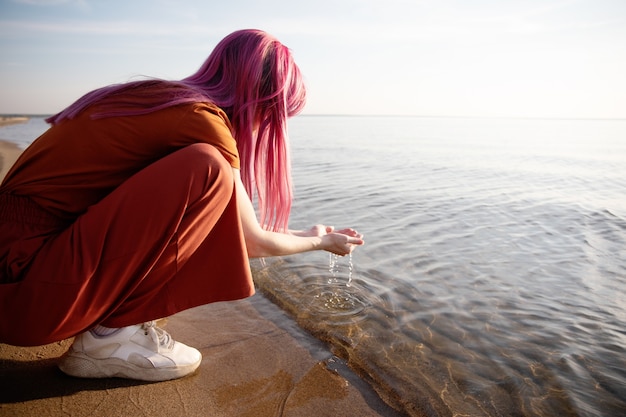 una donna con i capelli rosa e i vestiti arancioni raccoglie l'acqua dal mare nei suoi palmi